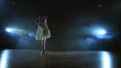 Ein-Junges-Mädchen-In-Einem-Weißen-Kleid-Tanzt-Ein-Modernes-Ballett,-Macht-Drehungen-Und-Sprünge-In-Zeitlupe-Auf-Der-Bühne,-Im-Hintergrund-Ist-In-Voller-Aufnahme-Rauch-Im-Scheinwerferlicht-Zu-Sehen.
